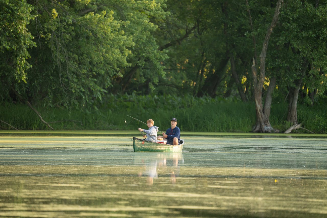 A father and son fish together in a canoe on a peaceful lake surrounded by lush greenery.